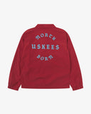 Back view flat shot of Uskees bright red polyester coach's jacket for men showing fgrahic text on the back in blue. Presented on a grey background.