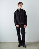 A male model wearing an Uskees black zip front jacket over a dark plaid shirt and black trousers, standing against a plain light gray wall and concrete floor.