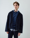 A man wearing a dark navy blue unzipped jacket over a textured blue sweater and striped shirt, standing against a plain light gray background.