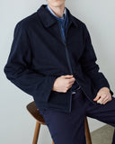A man shown neck to thigh wearing an Uskees dark navy blouson zip jacket over a blue knit sweater and white collared shirt, seated on a wooden stool against a light gray wall.