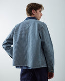 A man shown from the back wearing an Uskees  blue and white striped canvas utility jacket with a solid navy blue corduroy collar, blue check shirt and navy pants, photographed against a plain light gray studio background.