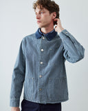 A man wearing an Uskees blue and white striped canvas utility jacket with a  contrasting dark blue collar, buttoned up, posed against a light gray studio background.