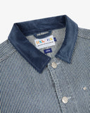 Front close up view of an Uskees blue and white striped canvas utility jacket showing the blue corduroy collar detail, silver coloured buttons and multi-coloured brand label.