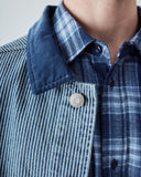 Close-up view of a model wearing an Uskees blue and white striped canvas utility jacket with a navy blue corduroy collar over a blue plaid shirt showing the collar detail.
