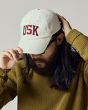 Model wearing cream Uskees 6-panel cap with 'USK' embroidered motif in maroon.