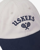 Close-up front view of cream and navy blue Uskees cap with 'USKEES' and tennis racquet motif embroidered in navy blue.
