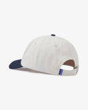 Side view of an Uskees cream and navy blue baseball cap showing the panels, the adjustable strap with metal fastener and a blue 'USKEES' logo tab.