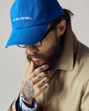 Model wearing bright blue Uskees 6-panel cap with 'All Will Be Well' embroidered motif in cream.