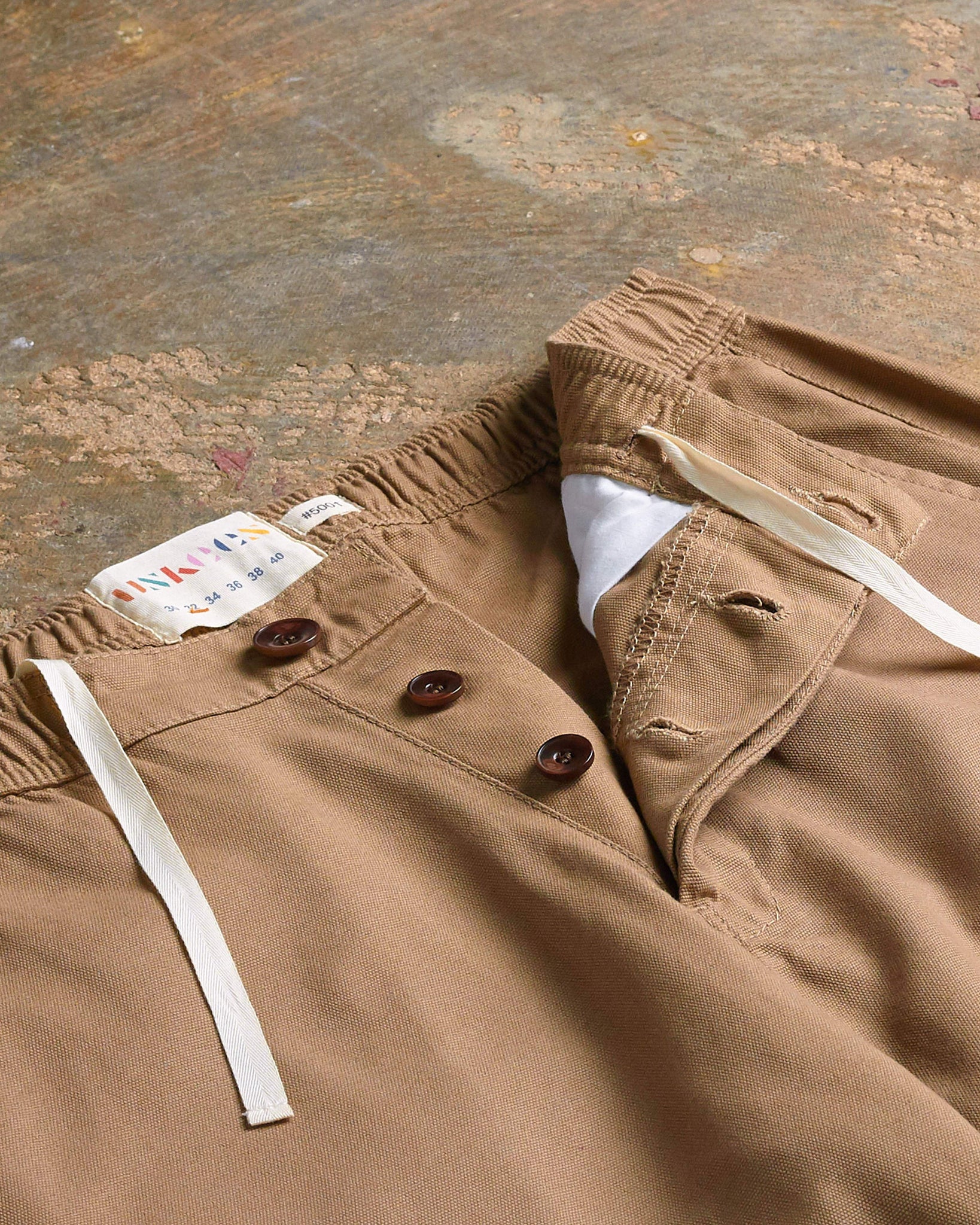 Flat and angled close-up view of Uskees khaki work pants with focus on natural corozo buttons for button fly and adjustable drawstring tie.