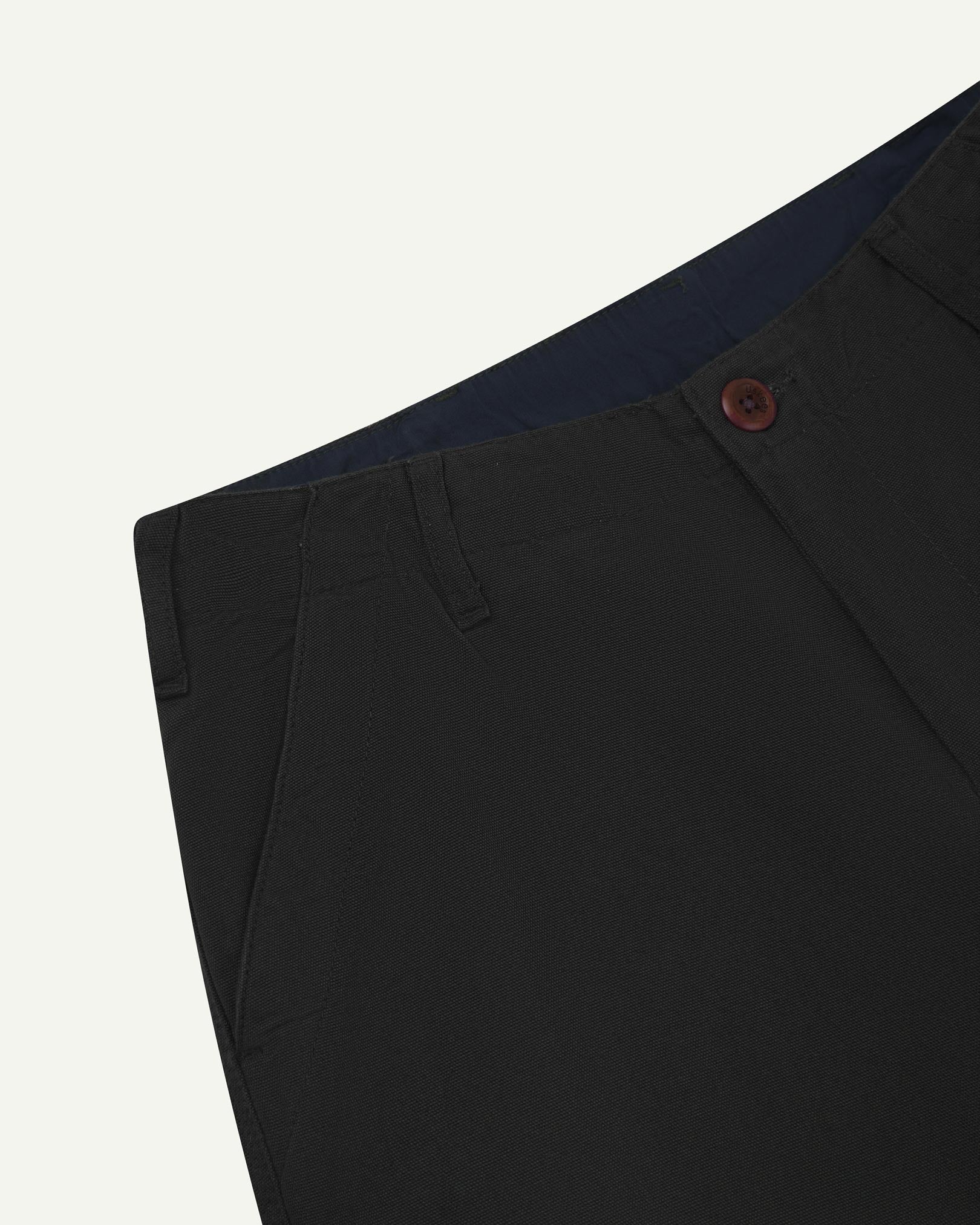 Close-up front view of left front pocket, belt loops, triple stitching, Corozo button and contrasting coloured lining material of black cotton pants.