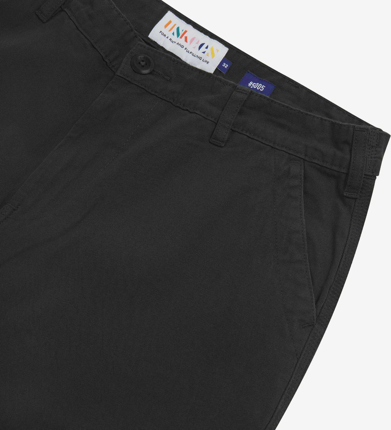 Close up of waist area of twill workwear pants in black. Clear view of front pocket, Corozo button fastening, Uskees branding label and organic cotton twill weave.