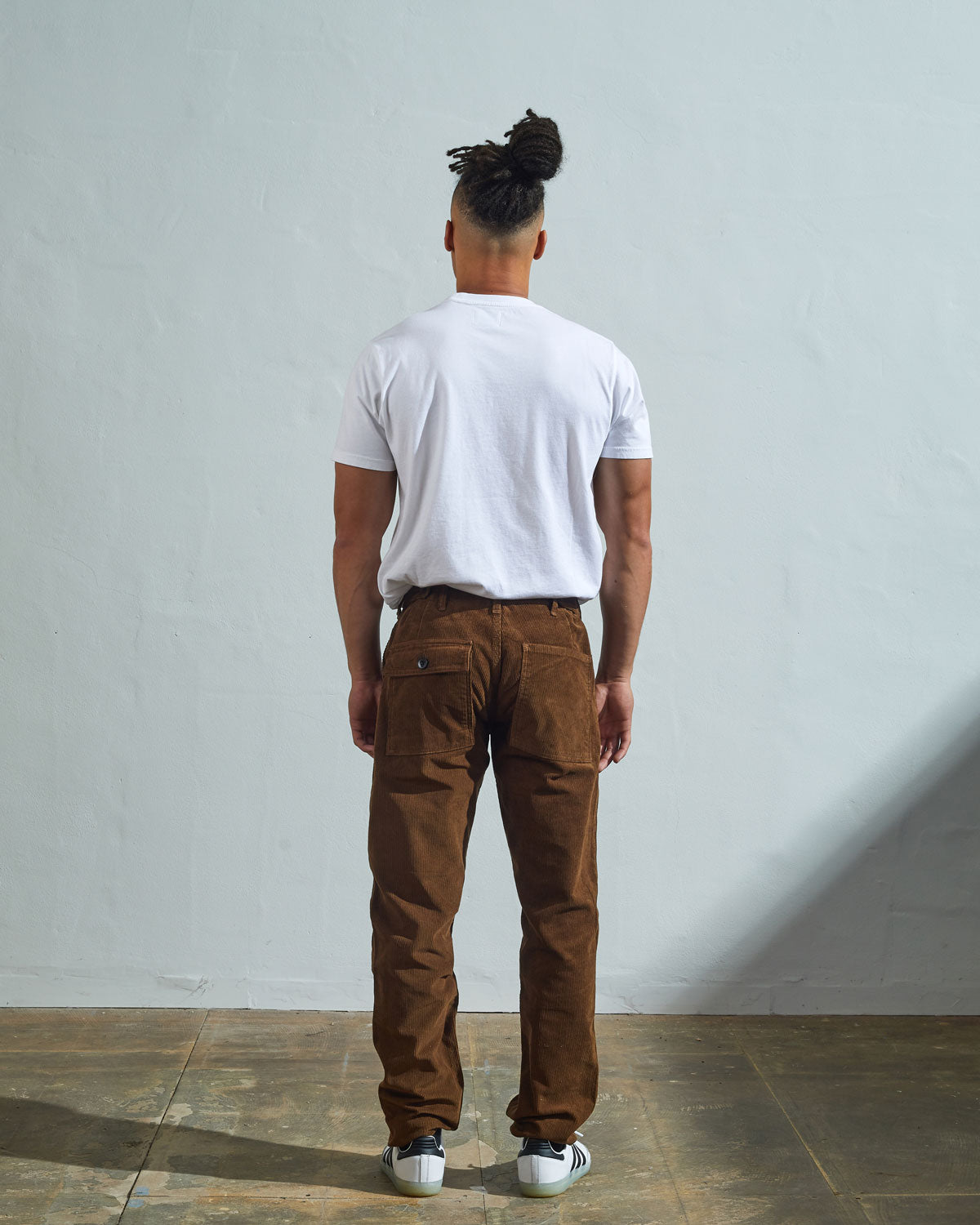 Full length back-view of model wearing brown corduroy 5005 trousers with view of rear pockets and belt loops. Paired with plain white t-shirt.