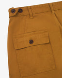 Close-up of Uskees mustard yellow men's trousers showing the waistband with two Corozo button adjusters and a  flap pocket with a matching button closure, on a light gray background.