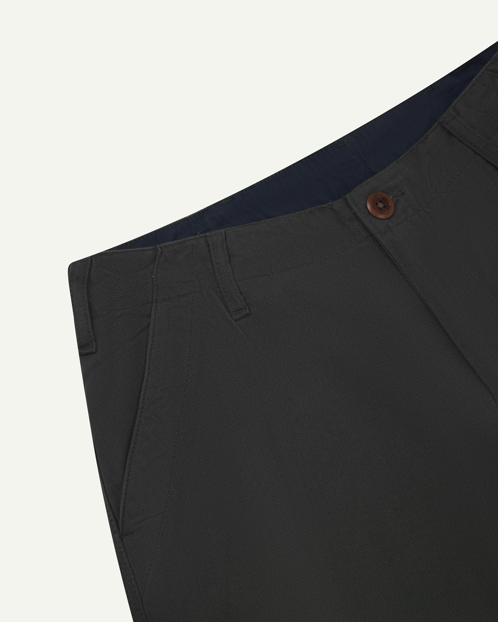 Close-up front view of left front pocket, belt loops, triple stitching, Corozo button and contrasting coloured lining material of charcoal-grey cotton pants.