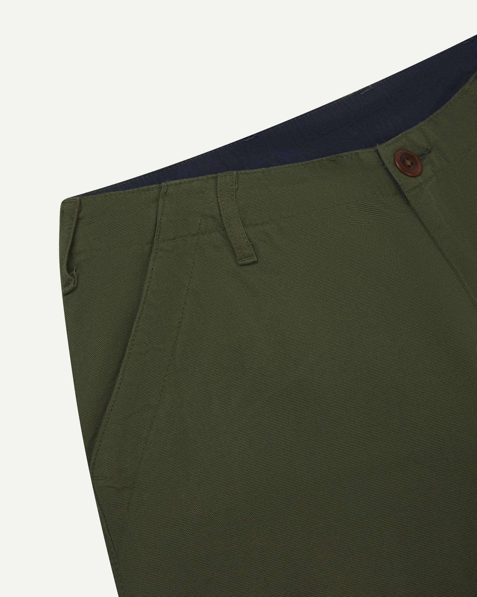 Close-up front view of left front pocket, belt loops, triple stitching, Corozo button and contrasting coloured lining material of coriander-green cotton pants.
