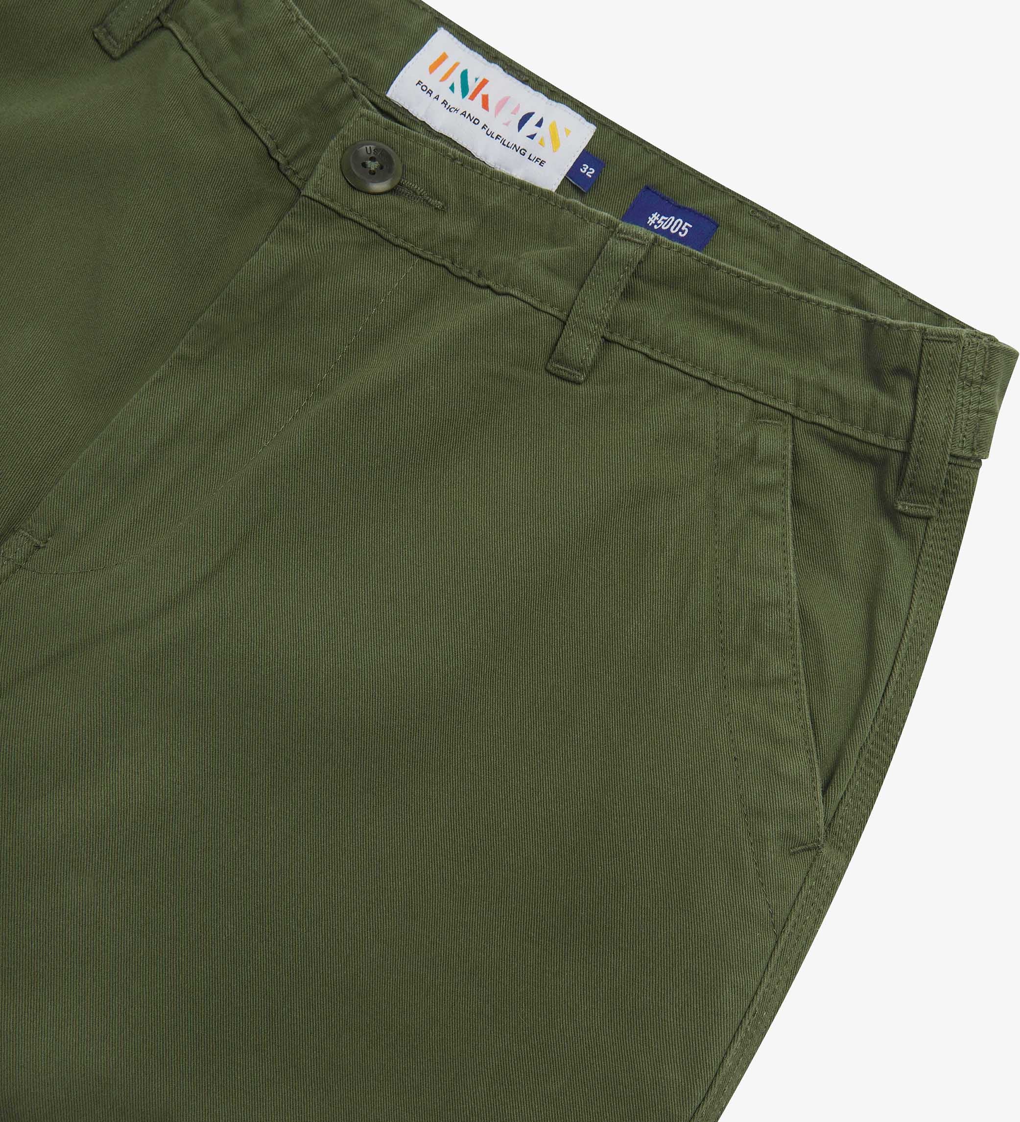 Close up of waist area of twill workwear pants in coriander-green. Clear view of front pocket, Corozo button fastening, Uskees branding label and organic cotton twill weave.