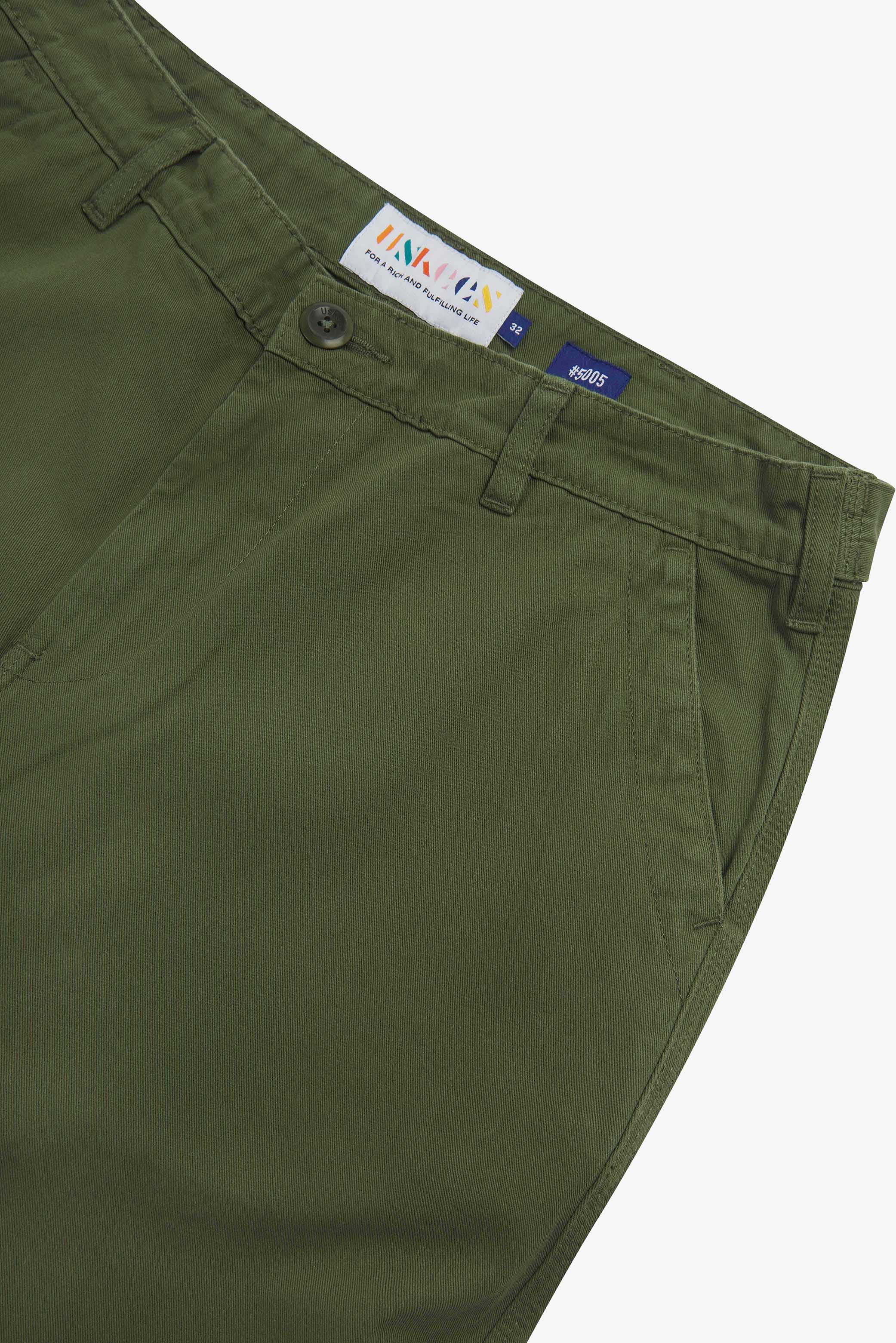 Close up of waist area of twill workwear pants in coriander-green. Clear view of front pocket, Corozo button fastening, Uskees branding label and organic cotton twill weave.