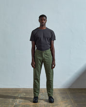 Full length front-view of model wearing coriander-green cotton 5005 workwear trousers with view of belt loops, corozo buttons and back pockets. Paired with faded black t-shirt.