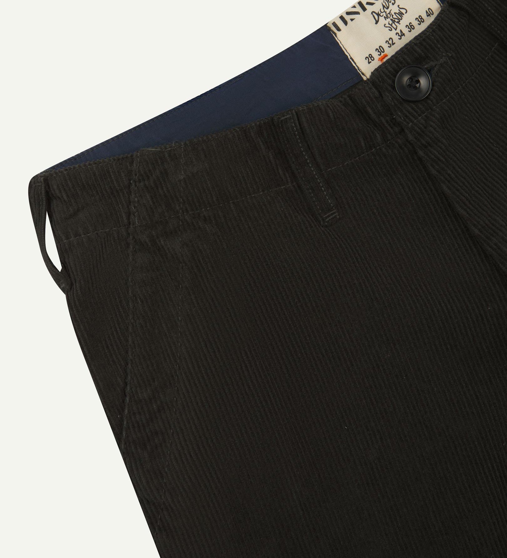 Close-up front view of left front pocket, belt loops, triple stitching, Corozo button and contrasting coloured lining material of faded black corduroy pants.