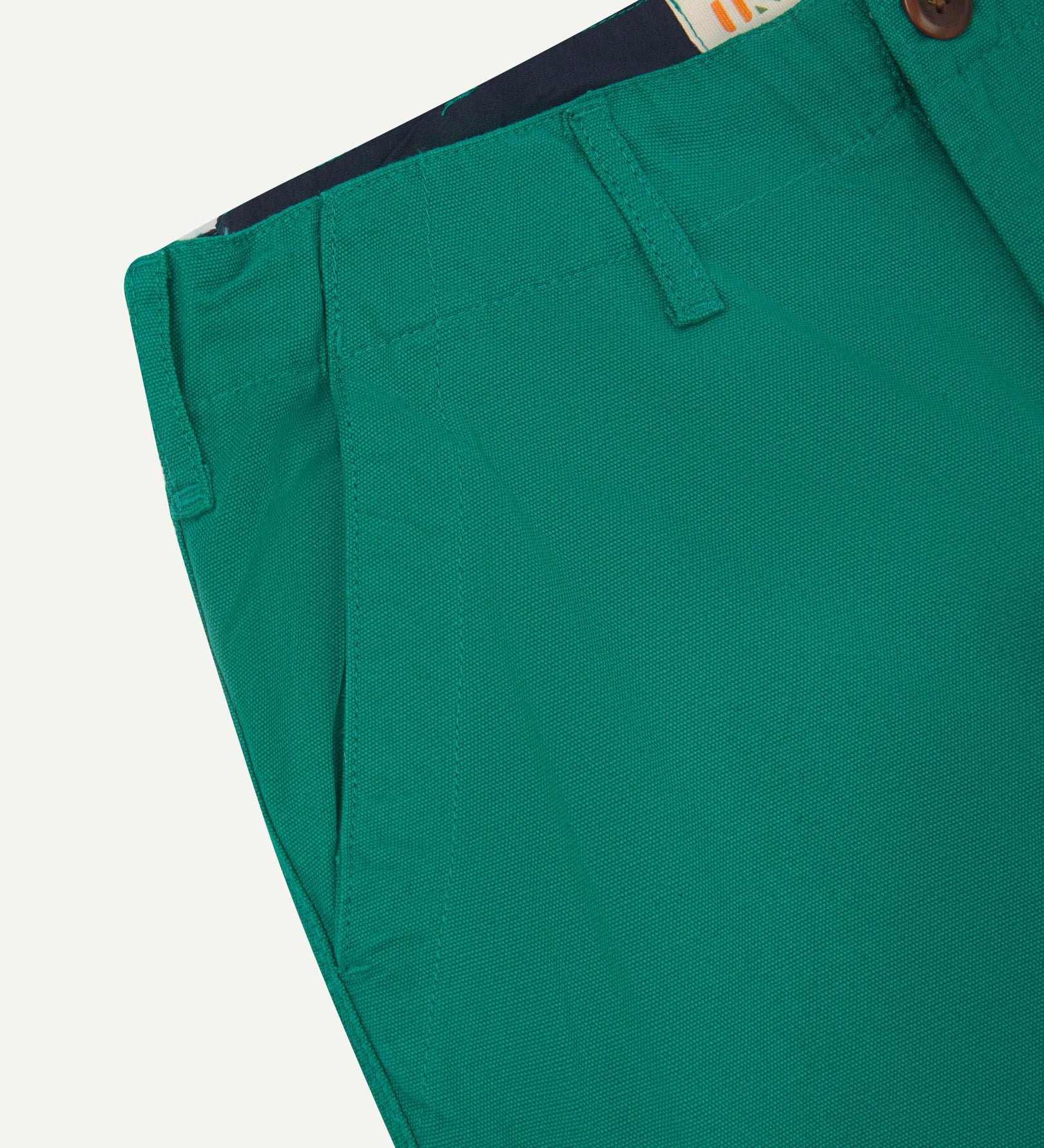 Close-up front view of left front pocket, belt loops, triple stitching, Corozo button and contrasting coloured lining material of foam green cotton pants.