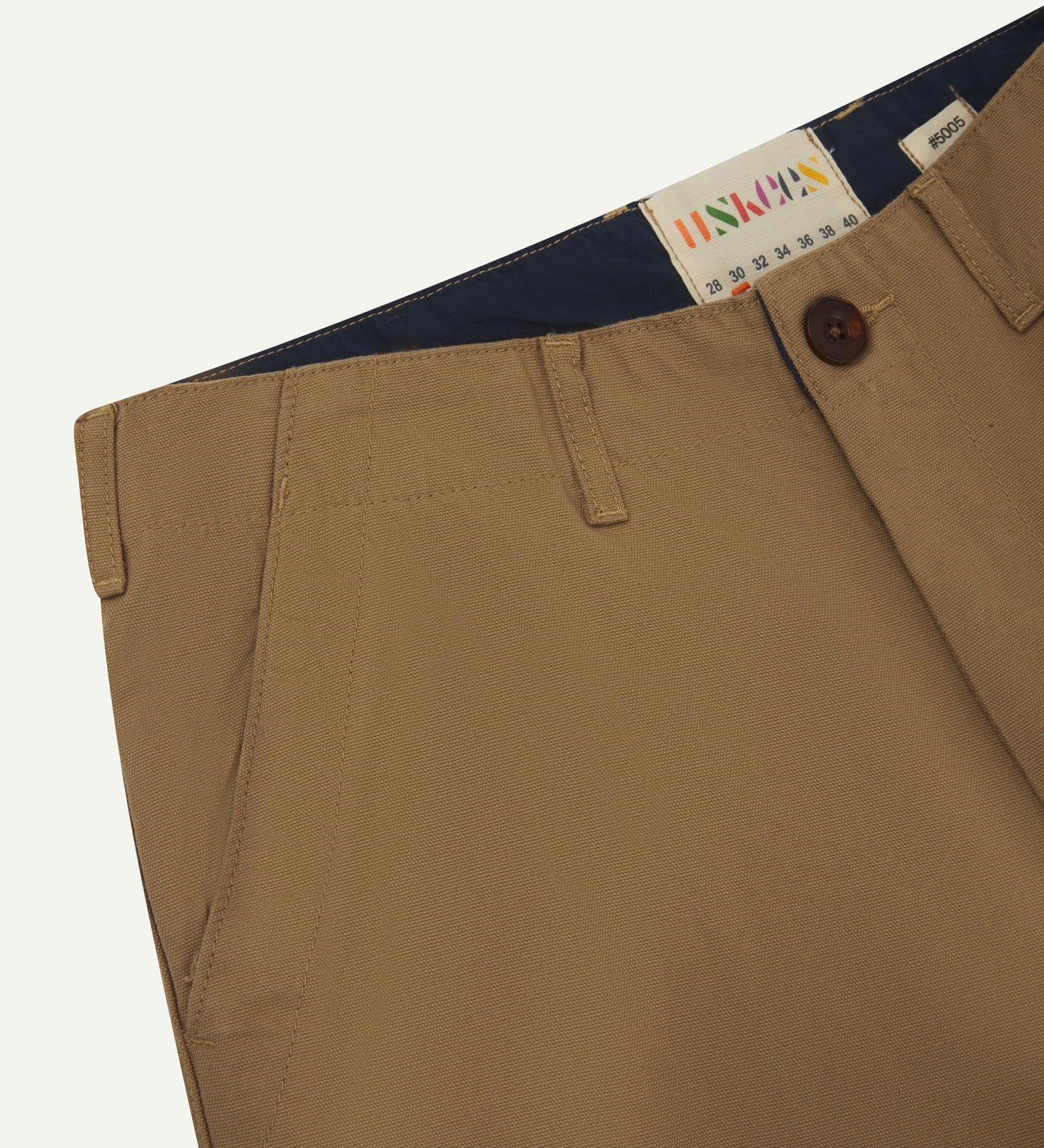 Close-up front view of left front pocket, belt loops, triple stitching, Corozo button and contrasting coloured lining material of khaki cotton pants.