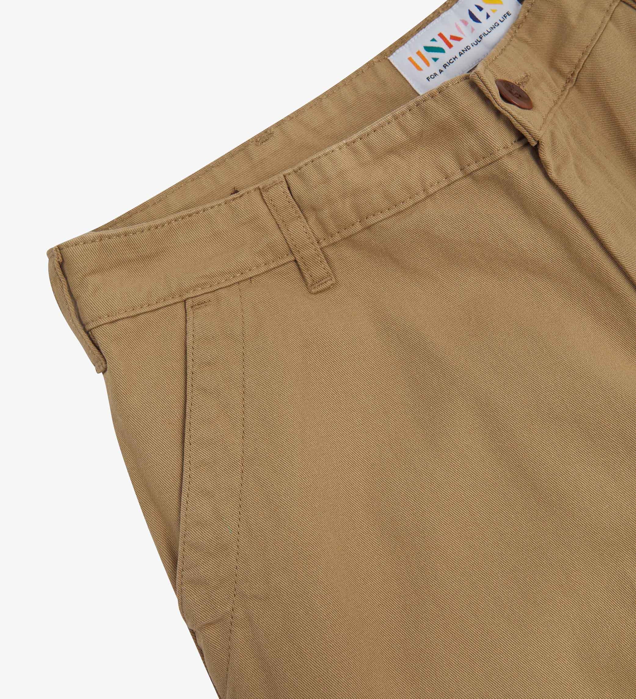 Close up of waist area of twill workwear pants in khaki. Clear view of front pocket, Corozo button fastening, Uskees branding label and organic cotton twill weave.