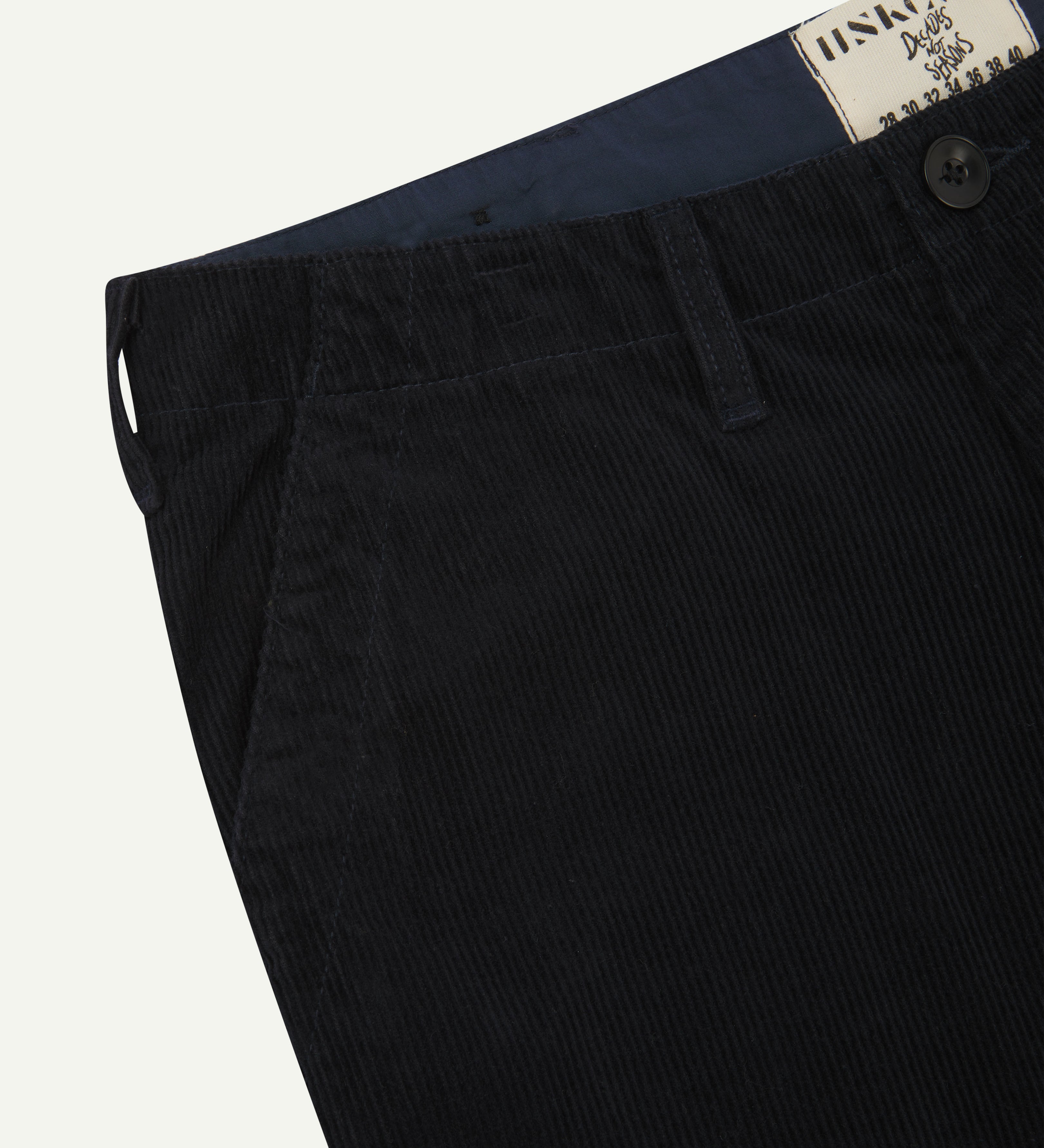Close-up front view of left front pocket, belt loops, triple stitching, Corozo button and contrasting coloured lining material of midnight blue corduroy pants.