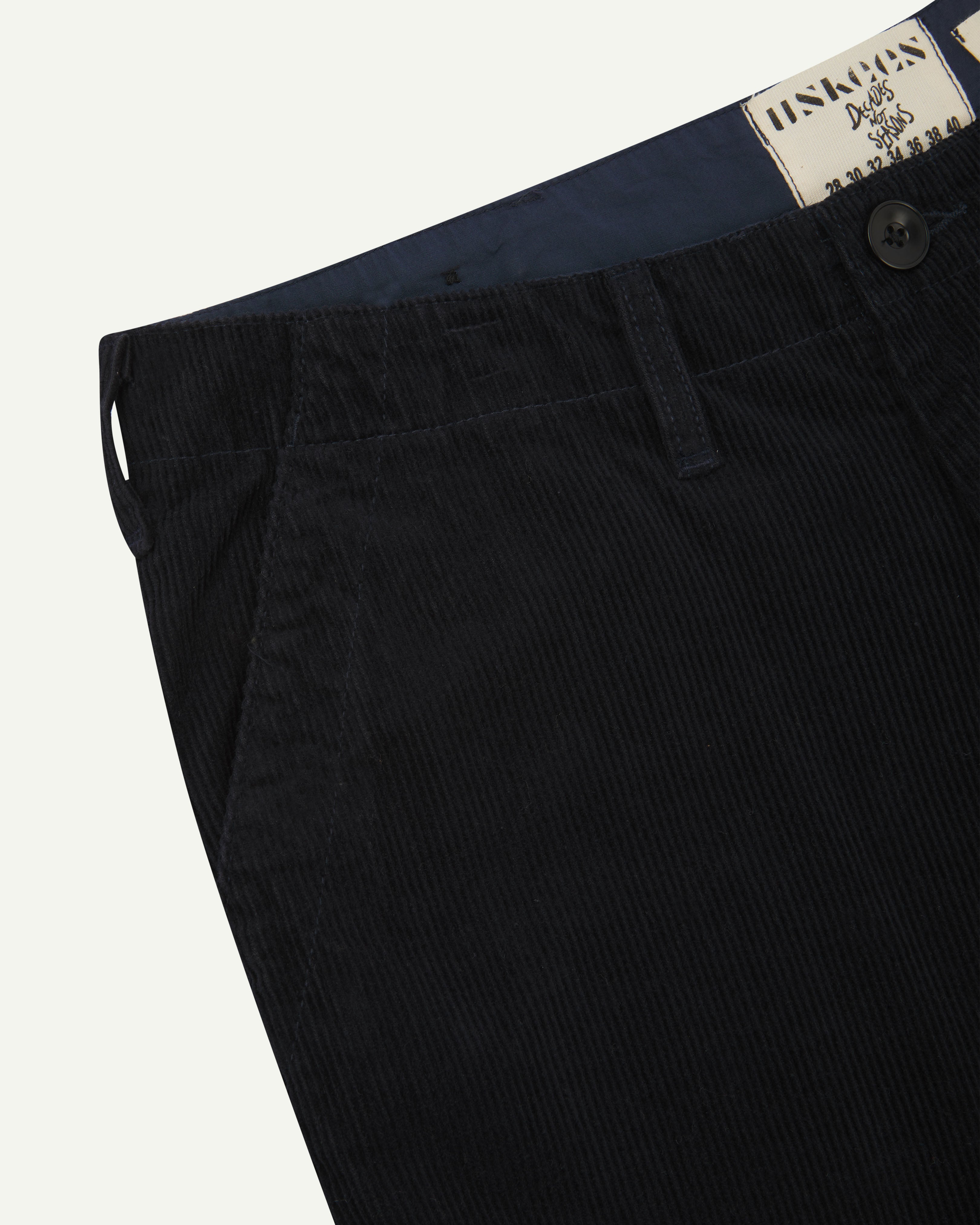 Close-up front view of left front pocket, belt loops, triple stitching, Corozo button and contrasting coloured lining material of midnight blue corduroy pants.