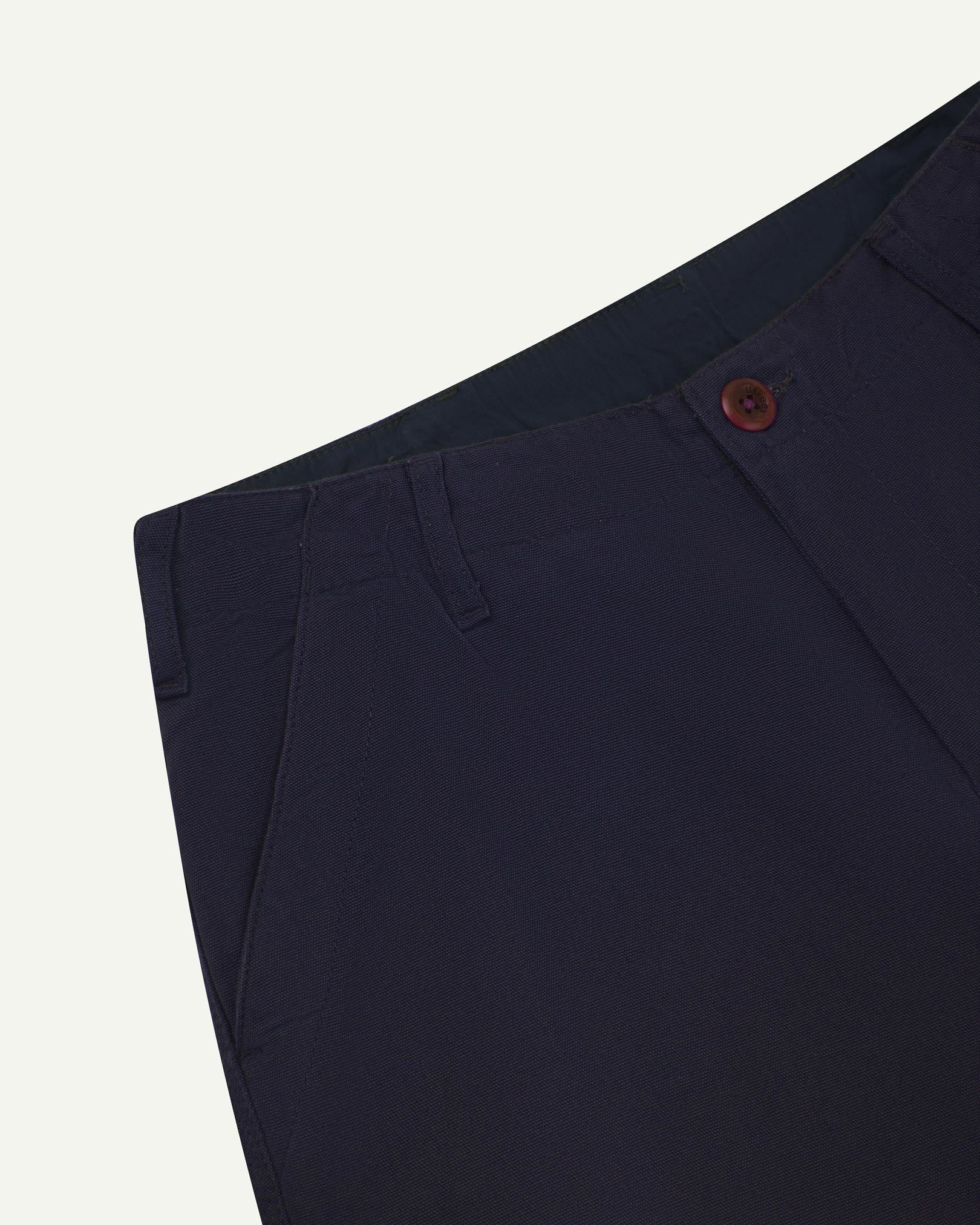 Close-up front view of left front pocket, belt loops, triple stitching, Corozo button and contrasting coloured lining material of midnight blue cotton pants.