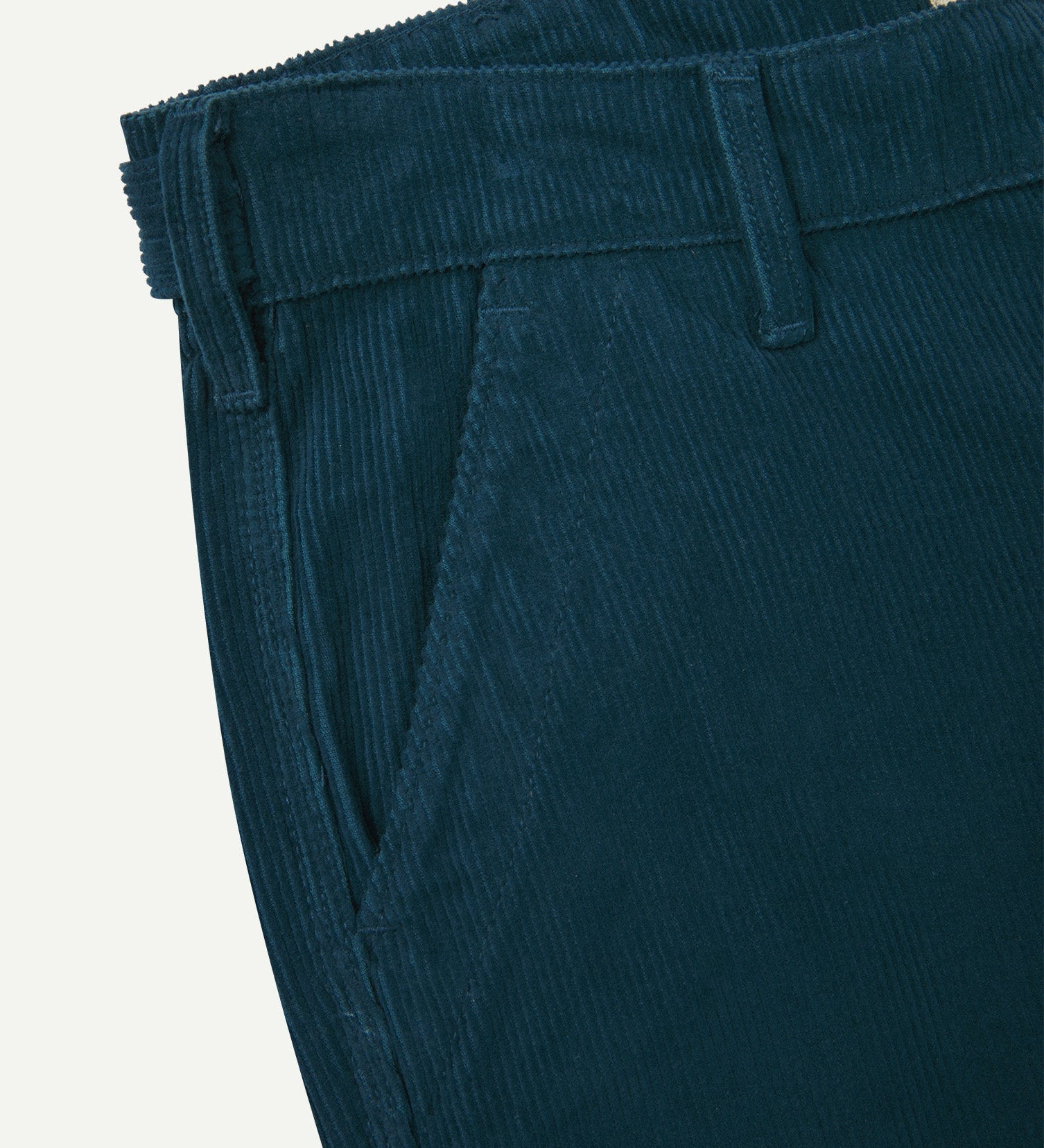 Close-up front view of left front pocket, belt loops, triple stitching, and Uskees branding label. Also a closer view of the organic, 11 wale corduroy in petrol blue.