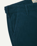 Close-up front view of left front pocket, belt loops, triple stitching, and Uskees branding label. Also a closer view of the organic, 11 wale corduroy in petrol blue.