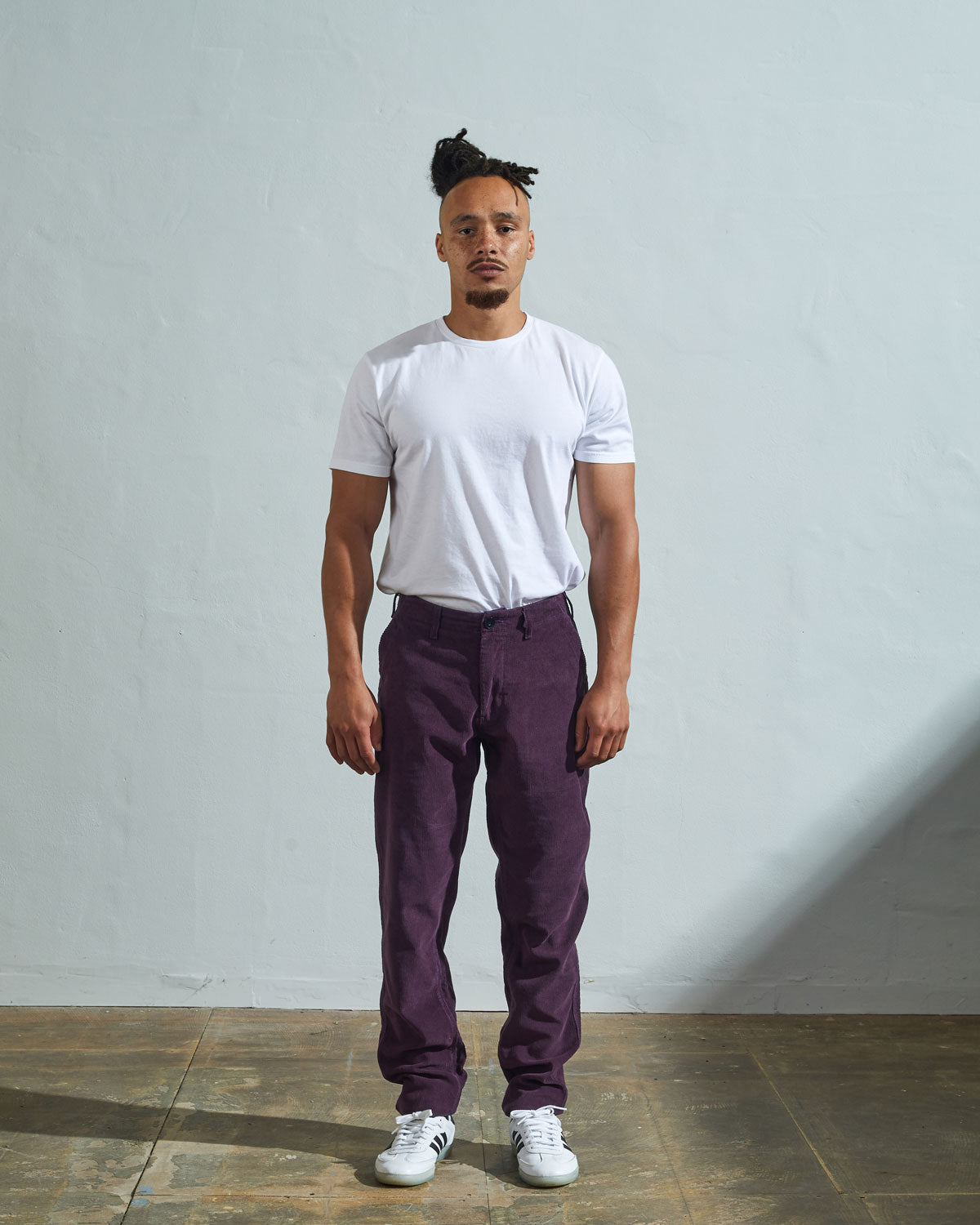 Full length front-view of model wearing purple-plum corduroy 5005 trousers with view of YKK zip fly and Corozo buttons. Paired with plain white t-shirt.