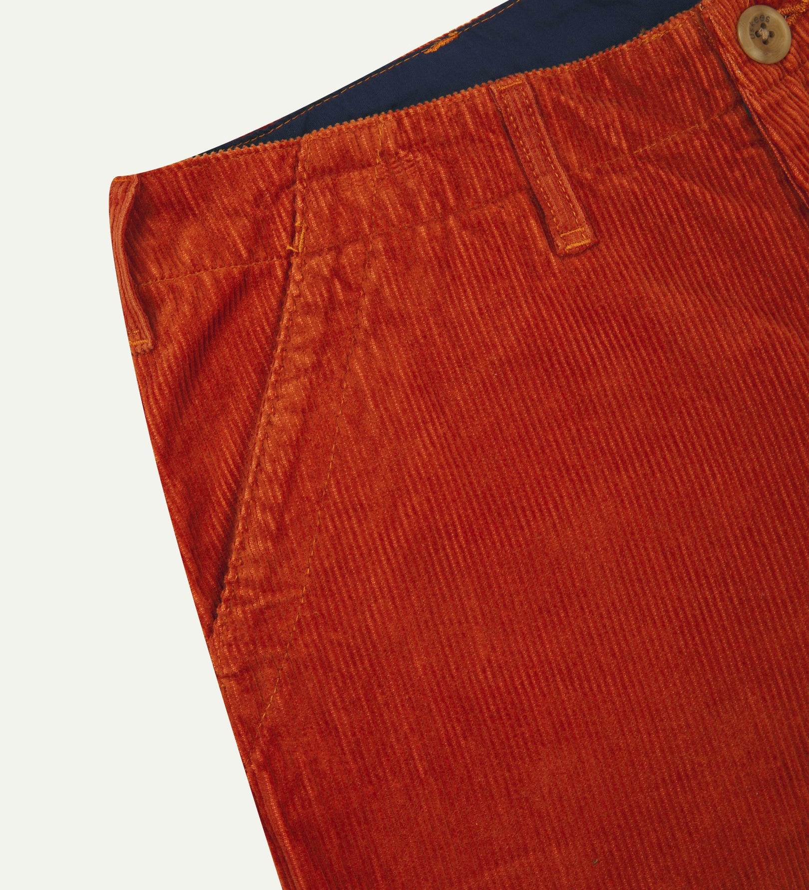 Close-up front view of left front pocket, belt loops, triple stitching, and contrasting lining. Also a closer view of the organic, 11 wale corduroy in rust-red.