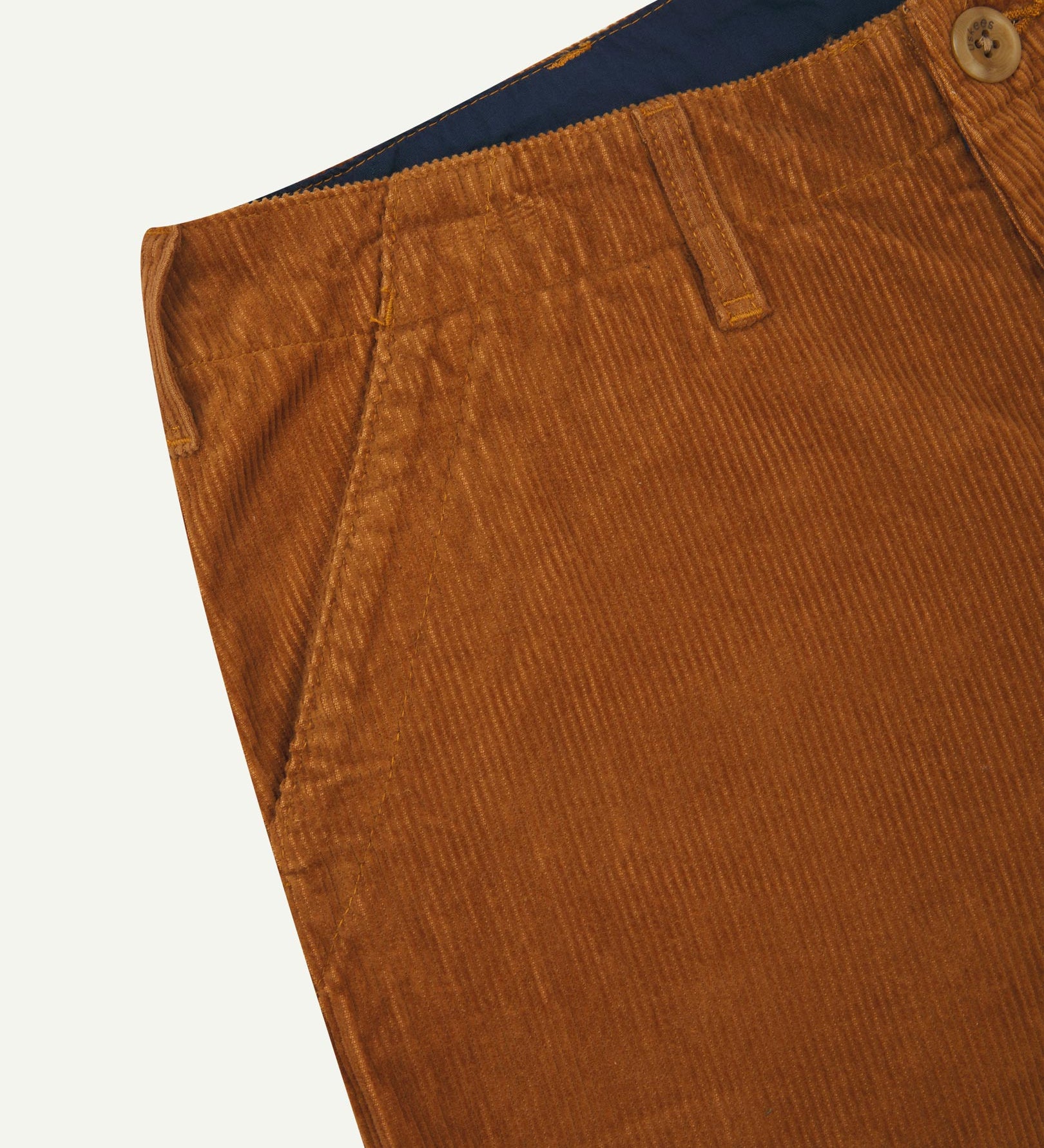 Close-up front view of left front pocket, belt loops, triple stitching, Corozo button and contrasting coloured lining material of tan corduroy pants.