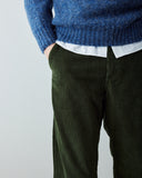 Close-up of a male model wearing Uskees dark green corduroy trousers, a blue knit sweater, and a white shirt, with one hand in the trouser pocket, against a plain light gray background.