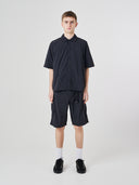 Full-length front view of a male model wearing an Uskees black short-sleeve button front trek shirt paired with matching Uskees technical shorts.