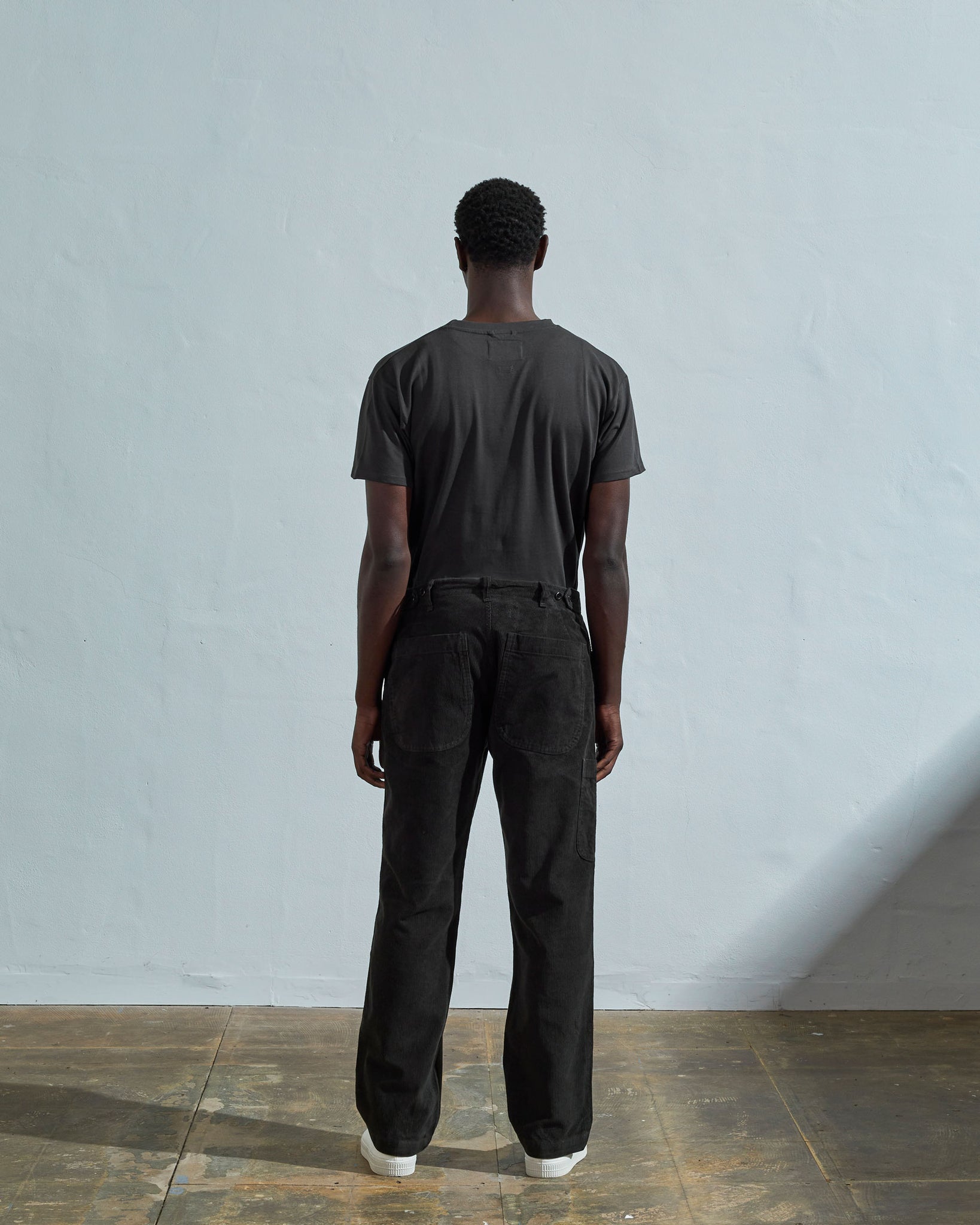 Full length reverse view of model wearing faded black corduroy 5012 trousers with view of straight leg fit, rear pockets and belt loops. Paired with matching t-shirt.