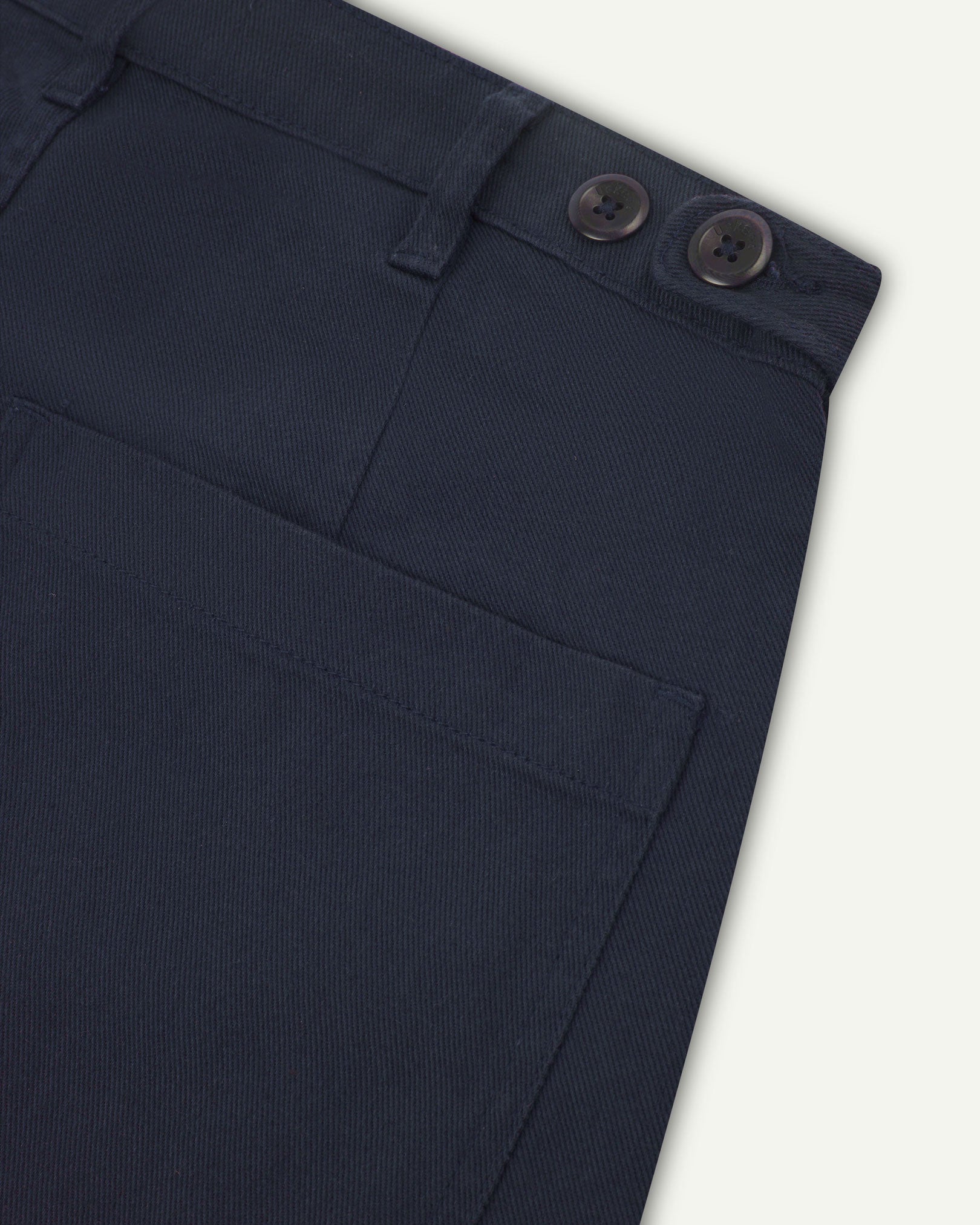 Reverse close-up of 5013 Uskees drill straight leg pants in dark blue, with focus on left rear pocket, belt loops and adjustable waistband with corozo buttons.