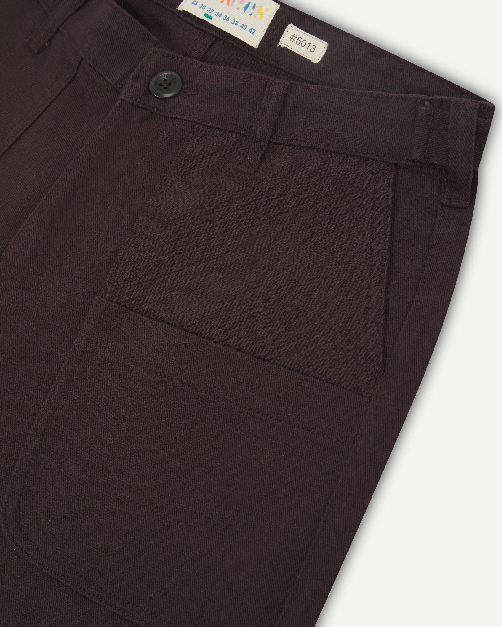 Front close-up of 5013 Uskees drill straight leg pants in brown-maroon, with focus on front pockets, layered pockets, belt loops waistband and Uskees branding label.