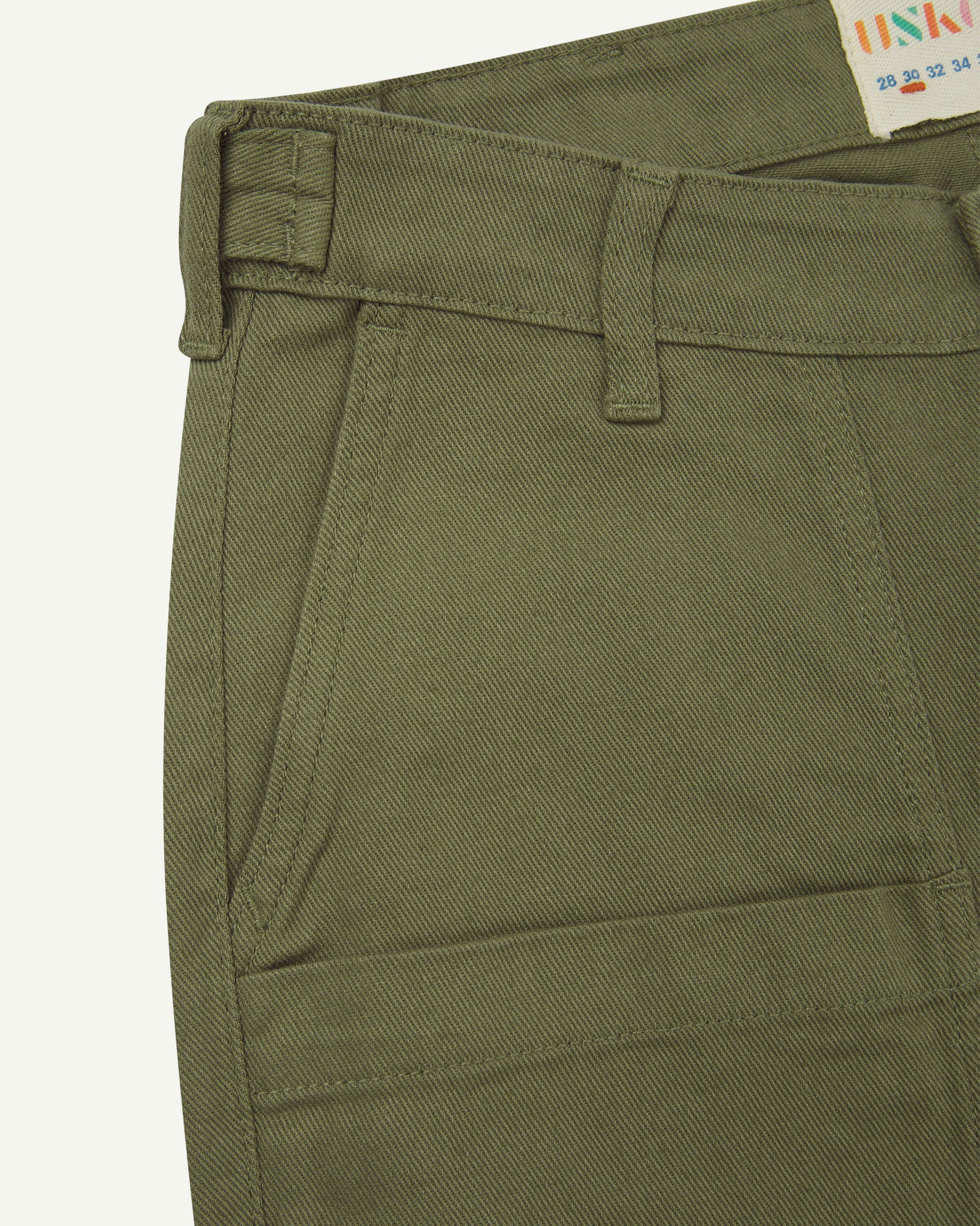 Front close-up of 5013 Uskees drill straight leg pants in moss green, with focus on front pockets, layered pockets, belt loops waistband and Uskees branding label.