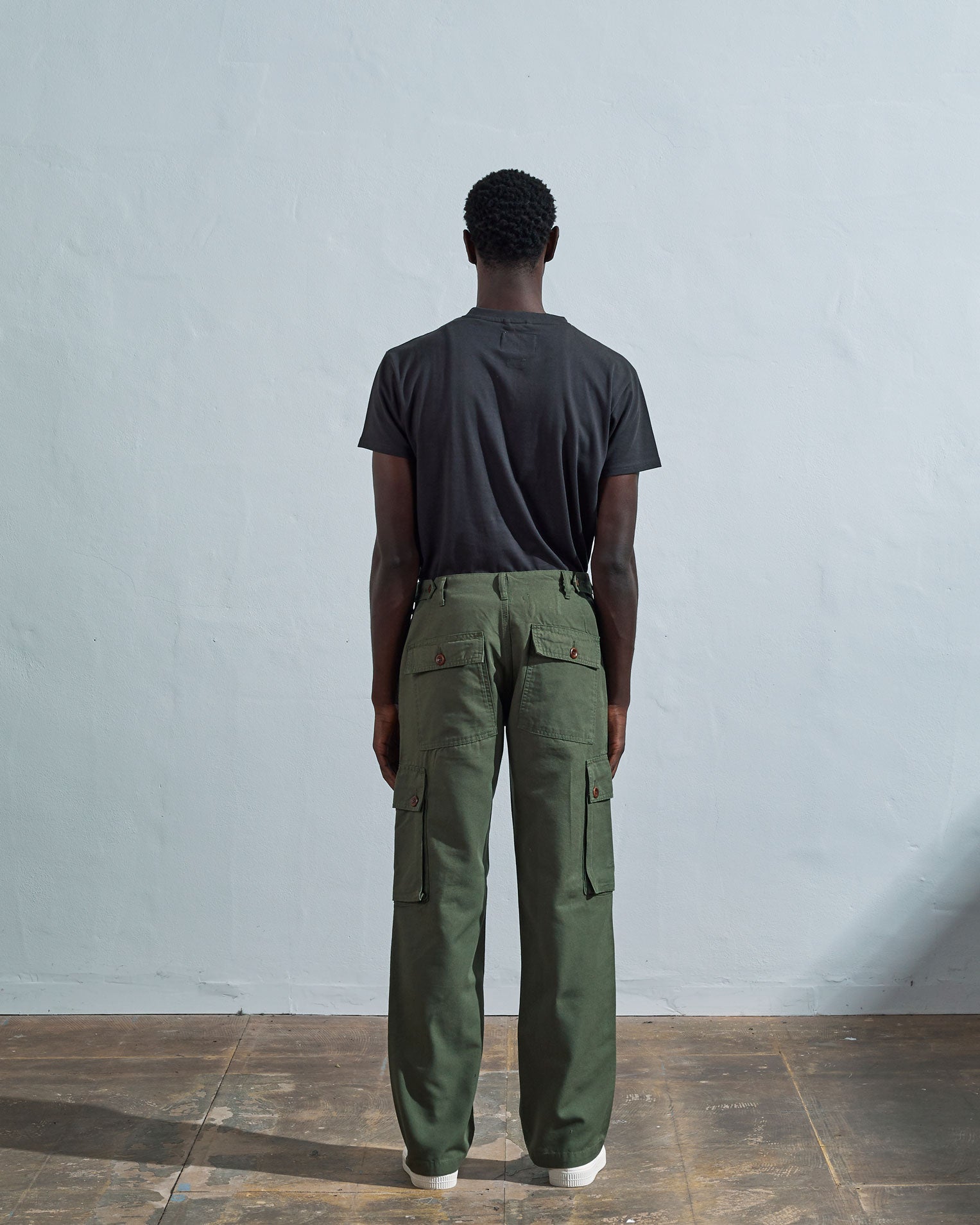 Full length back-view of model wearing coriander-green organic cotton #5014 trousers with view of rear and cargo pockets. Paired with faded black Uskees t-shirt.