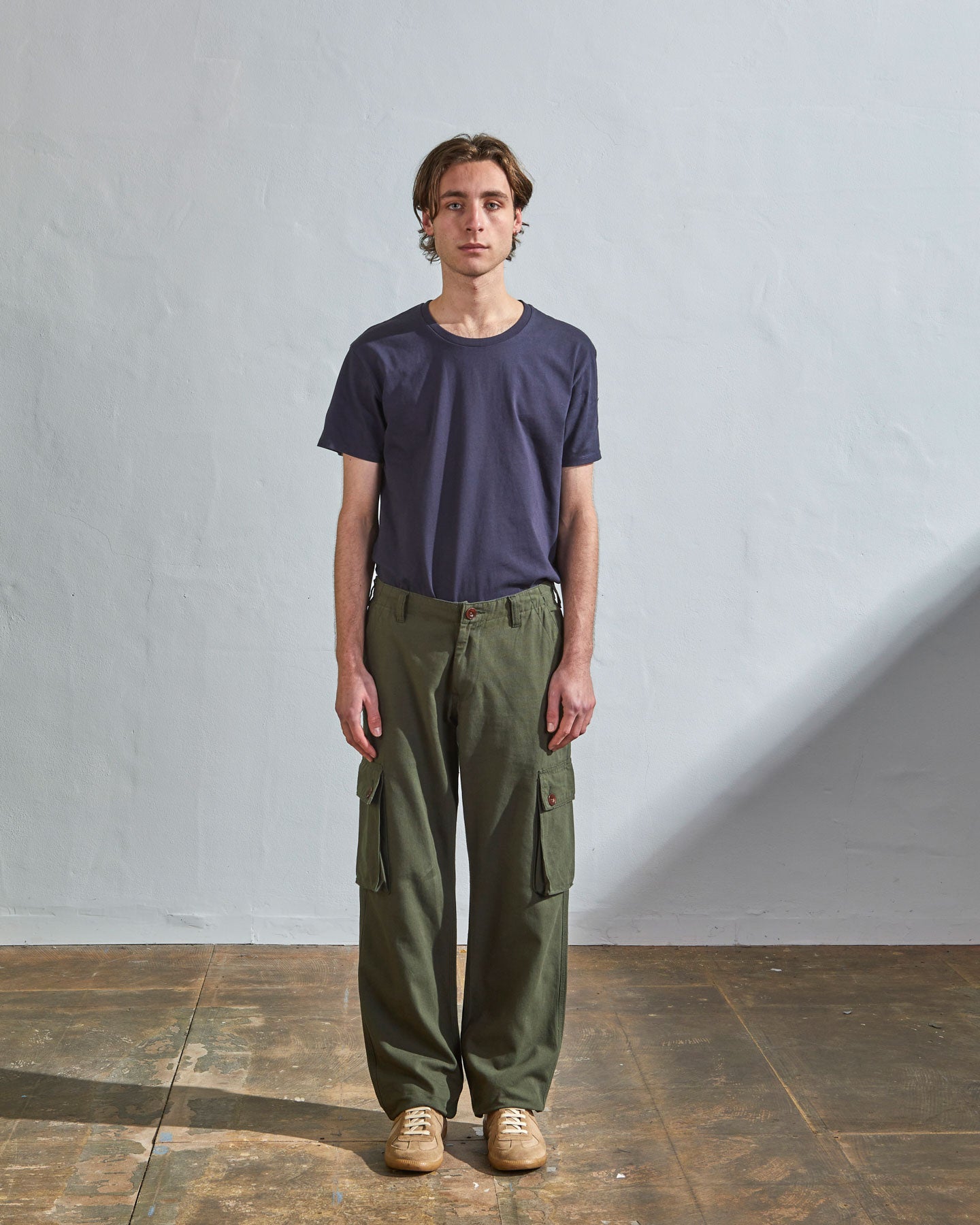 Full length front-view of model wearing vine green cotton 5014 trousers with view of belt loops, cargo pockets and Corozo buttons. Paired with midnight blue t-shirt.