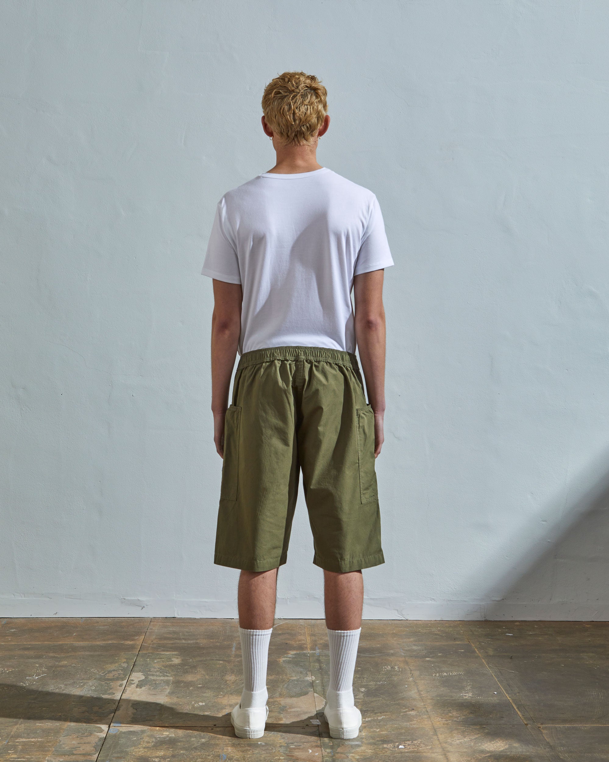 Back view of model wearing olive, loose leg light organic cotton shorts by Uskees. Large front pockets visible.