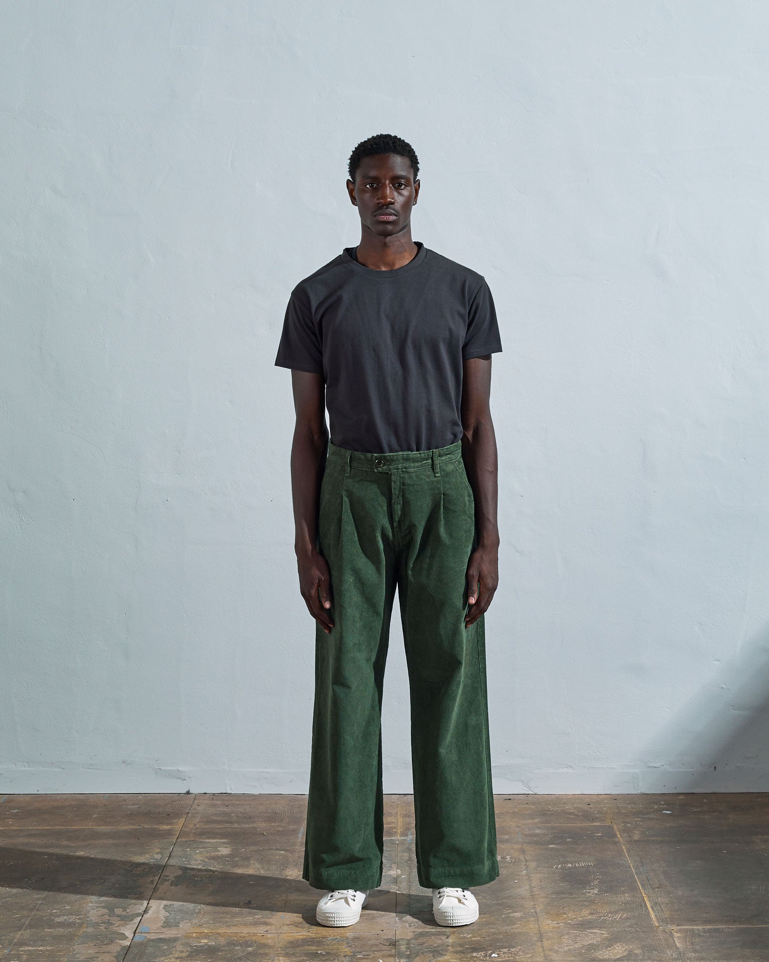 Full length front-view of model wearing coriander-green corduroy #5018 trousers with view of corozo button fastening. Paired with Uskees faded black t-shirt.