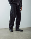 Male model seen from the waist down wearing Uskees black trousers, matching blazer, and black leather loafers, standing against a plain light gray wall and concrete floor.