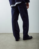 A male model shown from the back wearing dark navy blue relaxed-fit Uskees trousers, a striped shirt, and black leather shoes, standing on a concrete floor against a white wall.