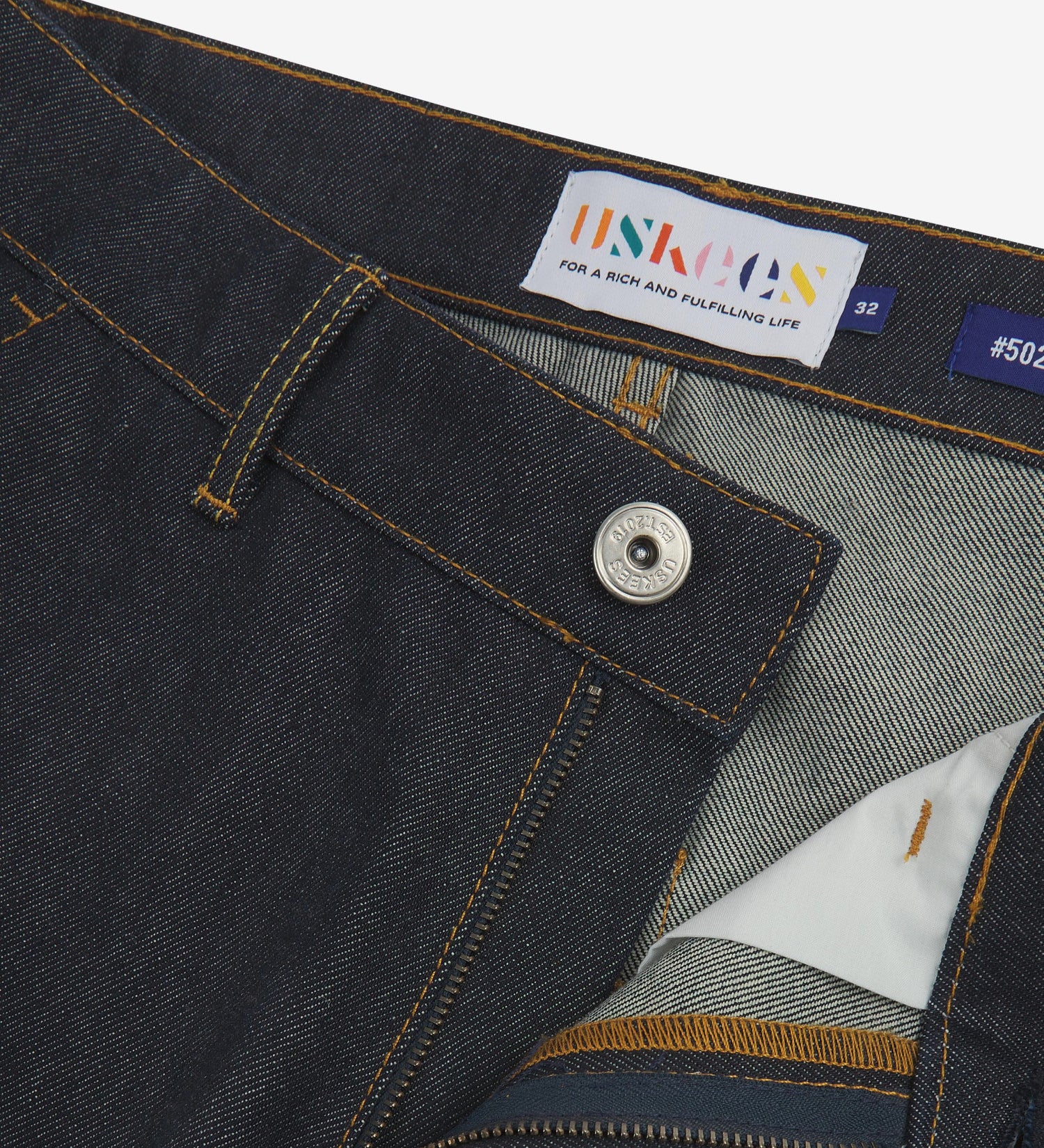 Close up of waist area of raw orta denim workwear pants in dark indigo. Clear view of front pocket, button fastening, Uskees branding label and clean denim weave.