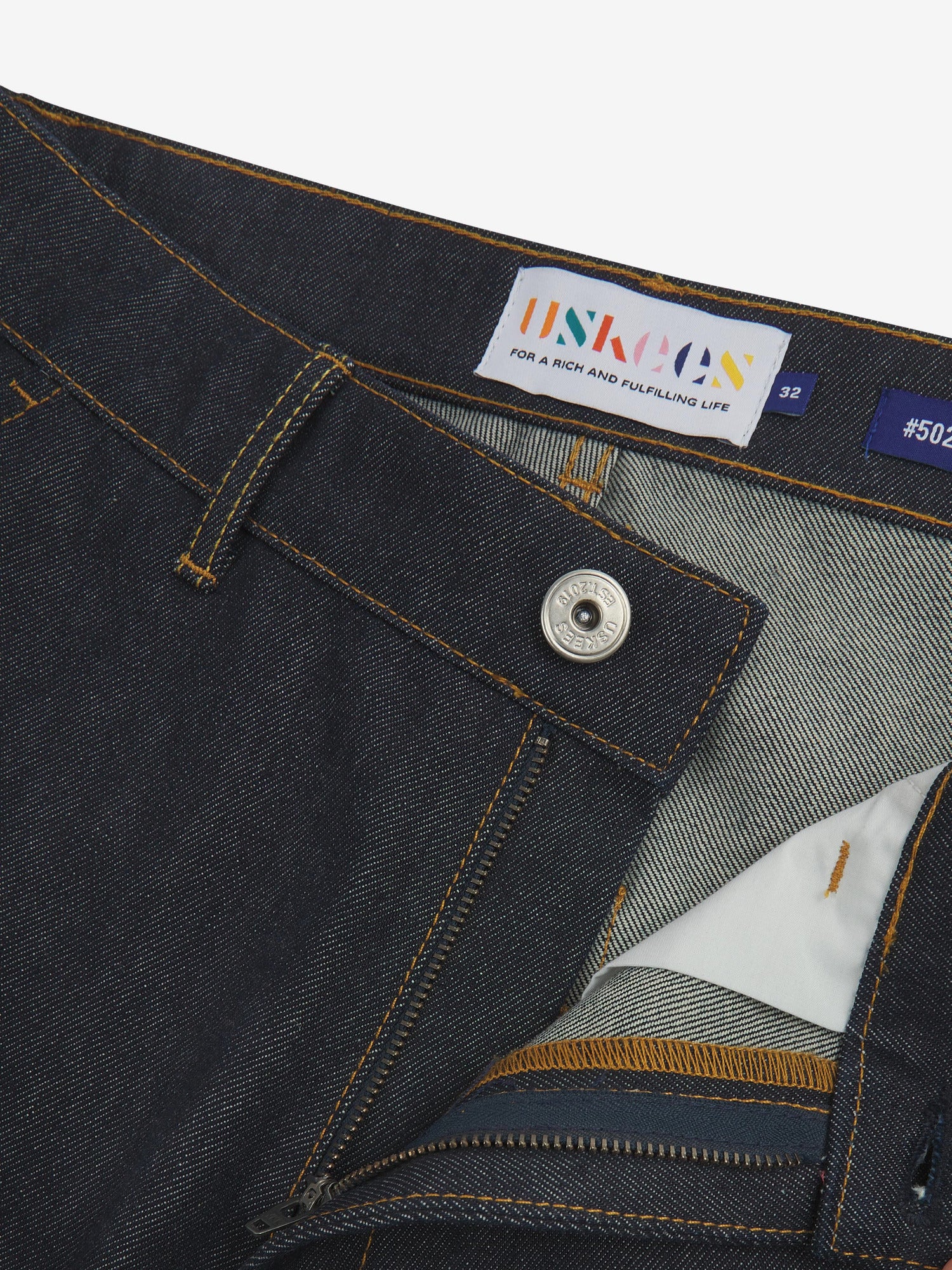 Close up of waist area of raw orta denim workwear pants in dark indigo. Clear view of front pocket, button fastening, Uskees branding label and clean denim weave.