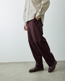Male model wearing Uskees dark burgundy loose fit trousers, a beige and olive green vertically striped button-down shirt, and dark brown leather loafers, standing against a plain light gray wall and concrete floor.
