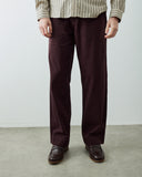 Male model wearing dark burgundy straight-leg trousers, a beige and dark green striped shirt, and dark brown leather loafers, standing against a plain light gray wall and concrete floor.